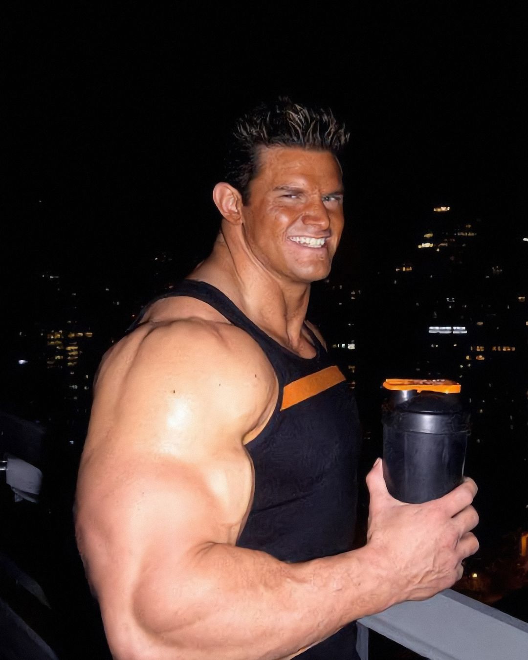 Elon Muskle grinning on a balcony at night with a protein shaker, city lights behind him